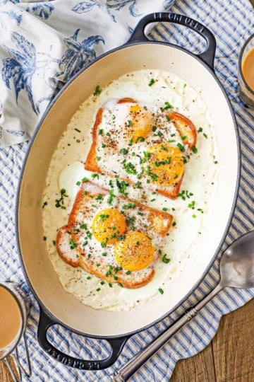 An overhead view of French Baked Eggs with Cream in an oval baking dish topped with snipped chives two glass mugs of coffee and a large silver serving spoon resting nearby.