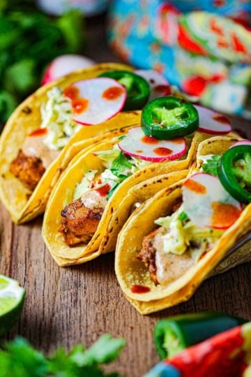 A straight-on view of three mahi mahi tacos sitting upright on a taco rack garnished with sliced radishes and fresh jalapeños.