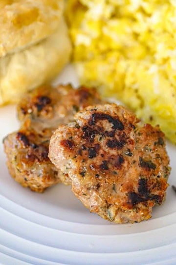 A close-up view of two homemade maple sausage patties on a white dinner plate next to a pile of scrambled eggs.