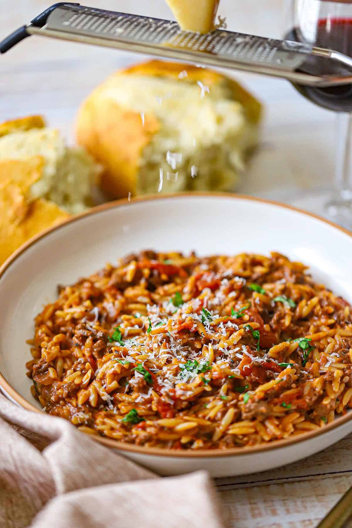A front-view of a shallow pasta bowl filled with a serving of One-Pot Ground Beef Orzo with a person grating fresh Parmesan cheese over the top.