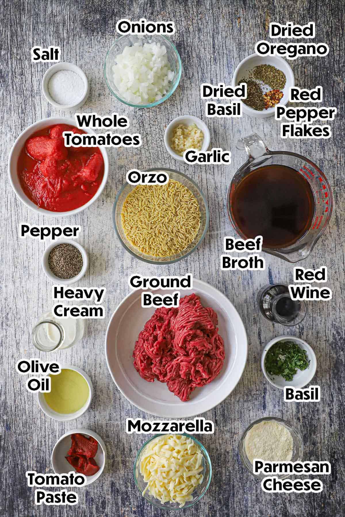 An arrangement of ingredients of One-Pot Ground Beef Orzo on a grey-wooden background including ground beef, dried orzo, whole tomatoes, onions, garlic, beef broth, cream, red wine, tomato, paste, cheese, spices, and herbs.