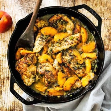 An overhead view of a large cast-iron skillet filled with pan-fried pork cutlets with peaches with a wooden spoon inserted off to the side.