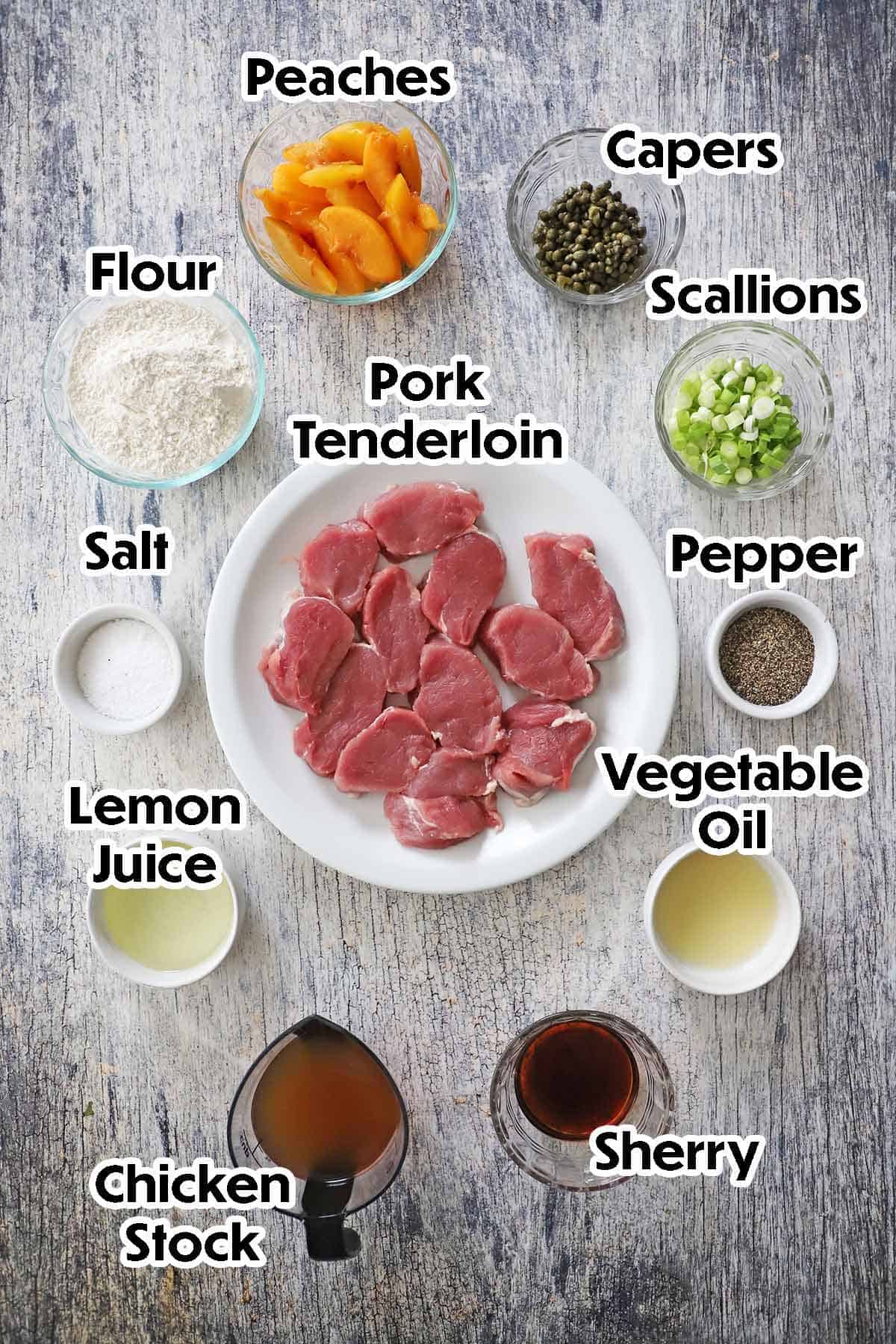 An arrangement of ingredients for pan-fried pork cutlets with peaches on a grey wooden background including sliced pork tenderloin, peaches, capers, scallions, flour, lemon juice, chicken stock, Sherry, oil, capers, and seasonings.