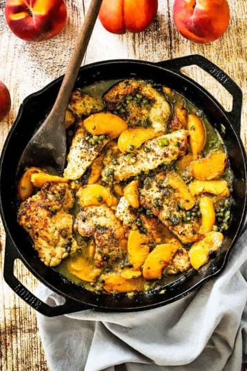 An overhead view of a large cast-iron skillet filled with pan-fried pork cutlets with peaches with a wooden spoon inserted off to the side and a few fresh peaches nearby.