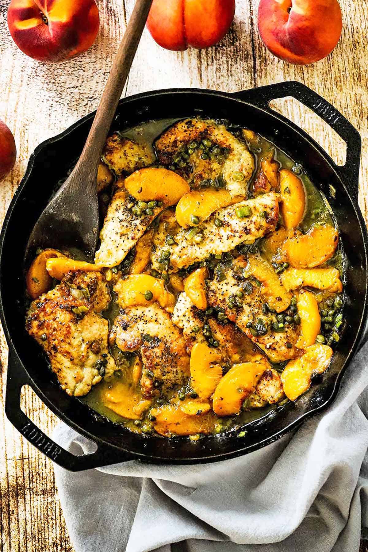 An overhead view of a large cast-iron skillet filled with pan-fried pork cutlets with peaches with a wooden spoon inserted off to the side and a few fresh peaches nearby.
