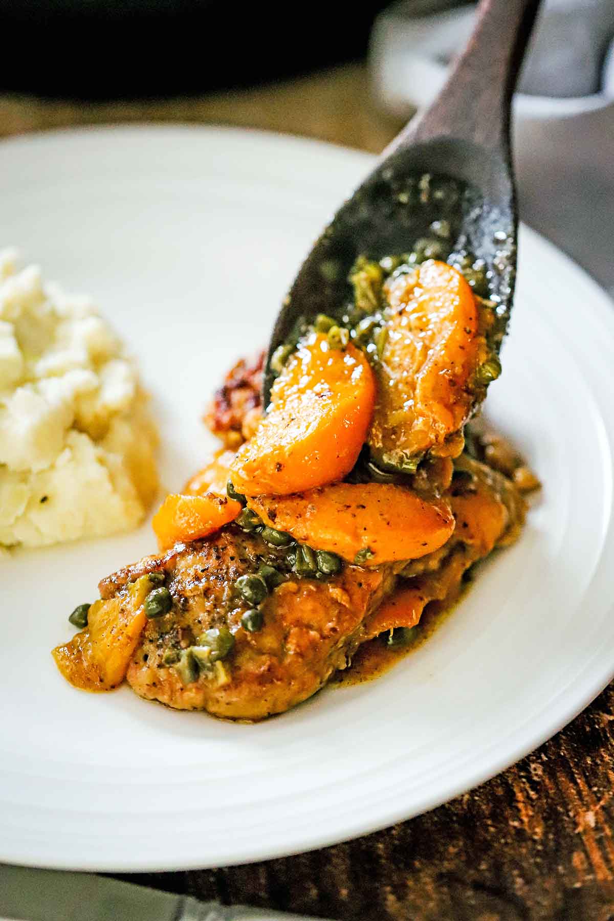 A large wooden spoon being used to place sautéed peaches and capers in a sauce over a pan-fried pork cutlet on a white dinner plate.