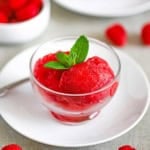 A close-up view of two scoops of Homemade Raspberry Sorbet in a small glass bowl on a white plate with a sprig of mint resting on top of the scoops of sorbet.