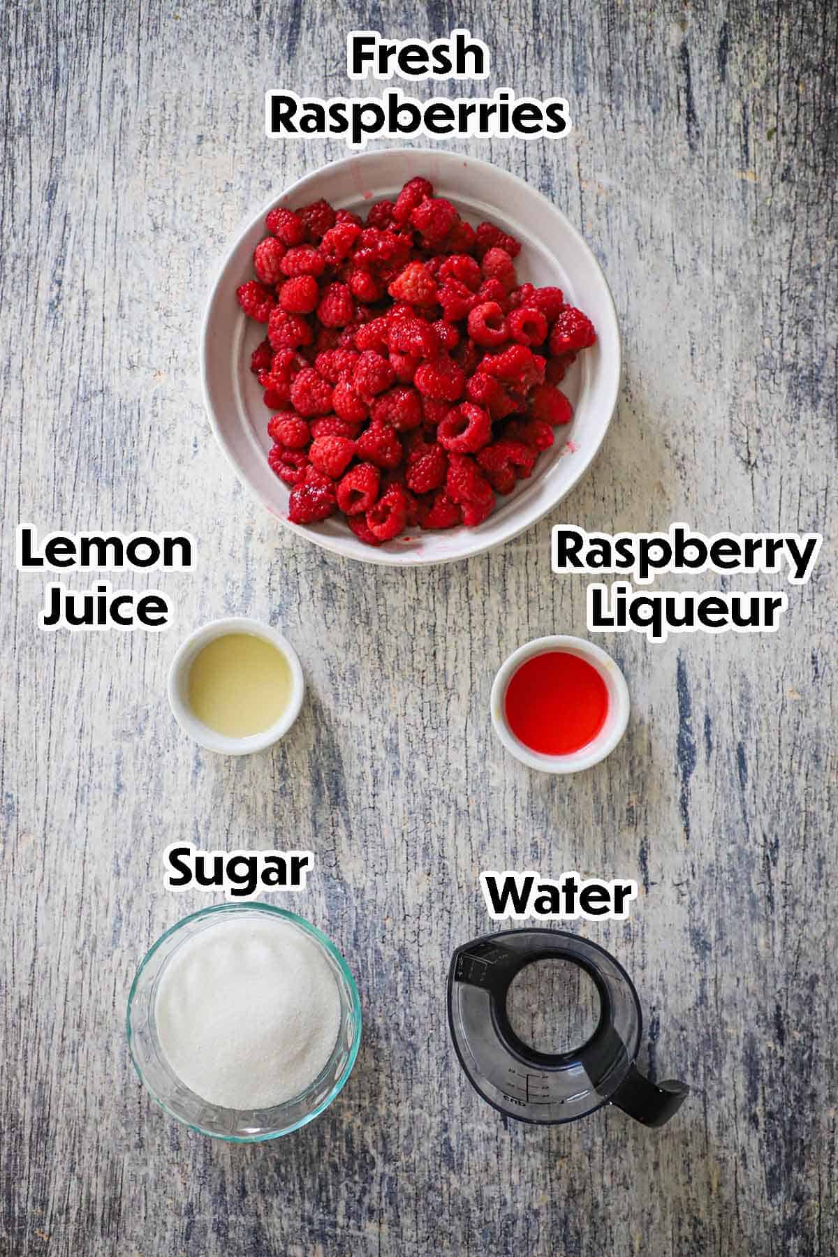 An arrangement of ingredients for homemade raspberry sorbet on a grey wooden background including fresh raspberries, lemon juice, sugar, water, and raspberry liqueur.