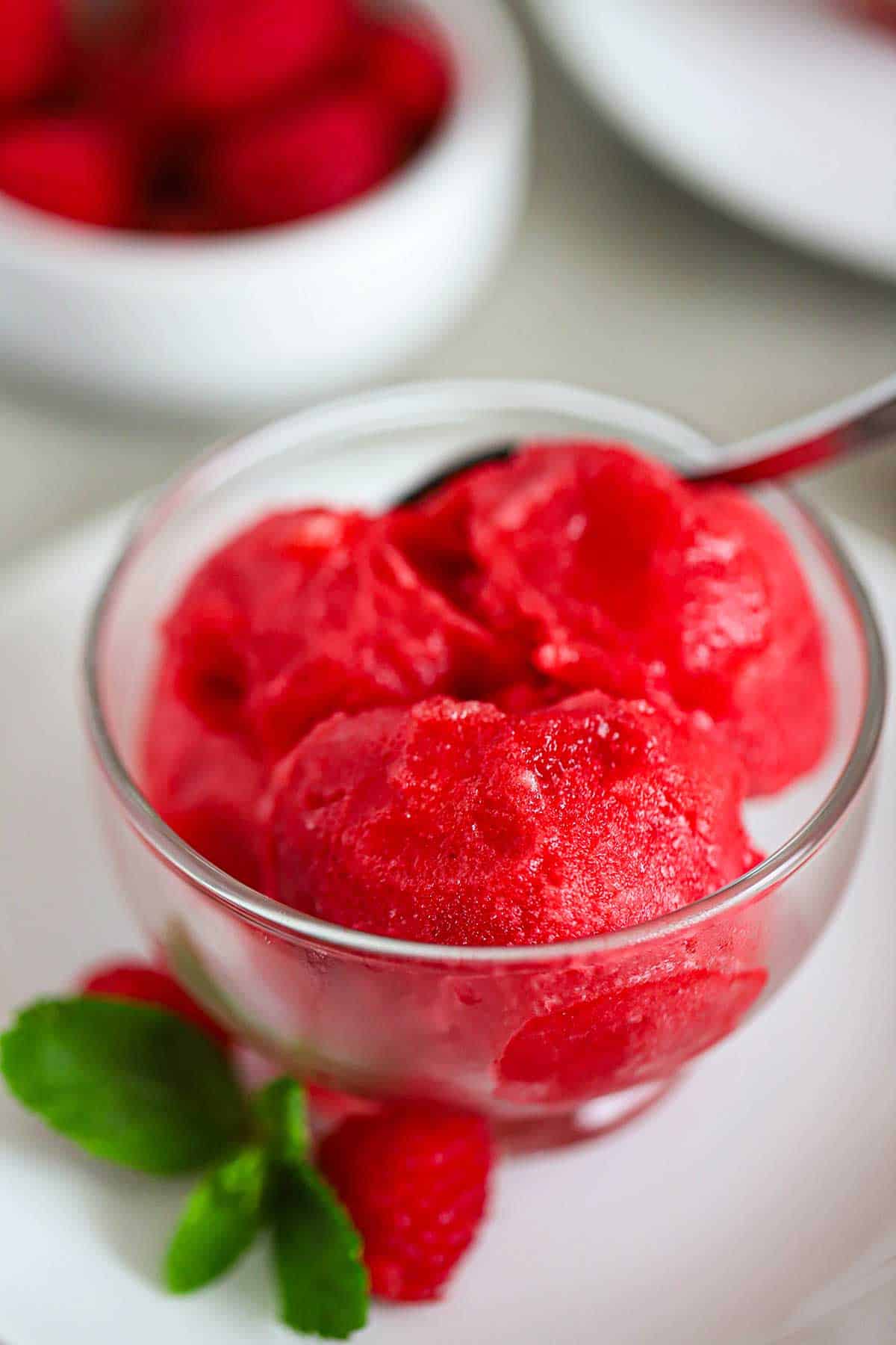A close-up view of three scoops of Homemade Raspberry Sorbet in a small glass bowl on a white plate with a sprig of mint and two raspberries sitting next to the bowl.