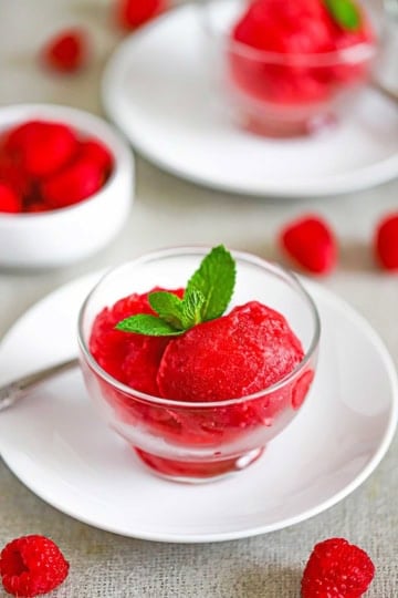 A close-up view of two scoops of Homemade Raspberry Sorbet in a small glass bowl on a white plate with a sprig of mint and two raspberries sitting next to the bowl.