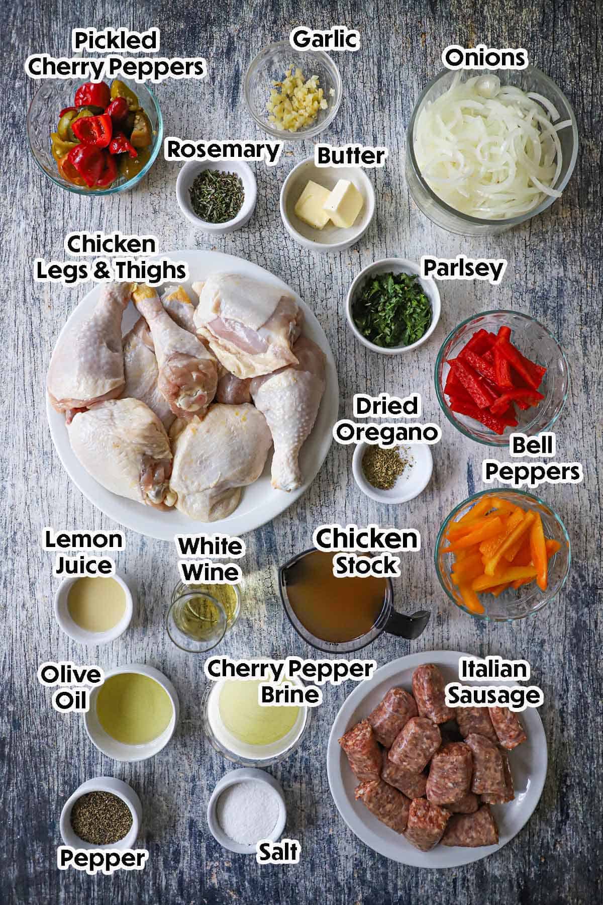 An arrangement of ingredients for chicken scarpariello on a grey wooden background including chicken legs and thighs, Italian sausage, bell peppers, onions, cherry pepers, garlic, chicken stock, wine, lemon juice, oil, butter, herbs, and seasonings.