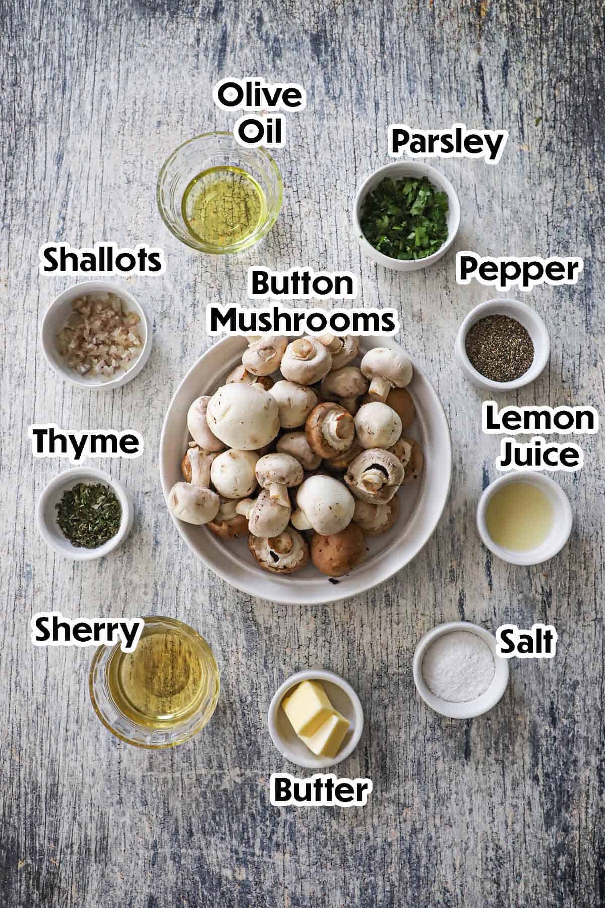 An arrangement of ingredients for sautéed mushrooms - steakhouse style on a grey wooden background including button mushrooms, shallots, olive oil, Sherry, butter, lemon juice, herbs, and seasonings.
