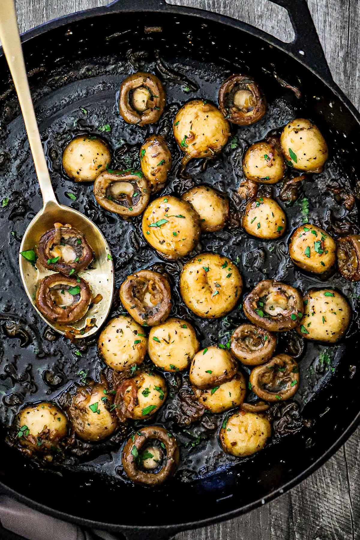 An overhead view of saut&eacute;ed mushrooms in a large black cast-iron skillet and garnished with chopped Italian parsley and a gold spoon resting in the skillet with a couple of mushrooms on it.