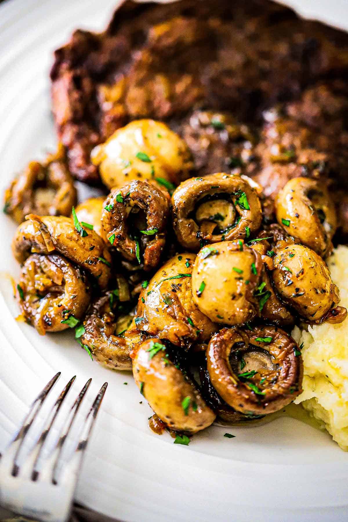 A straight-on view of a pile of sautéed mushrooms - steakhouse style on a white dinner plate next to a cooked steak and a pile of mashed potatoes.