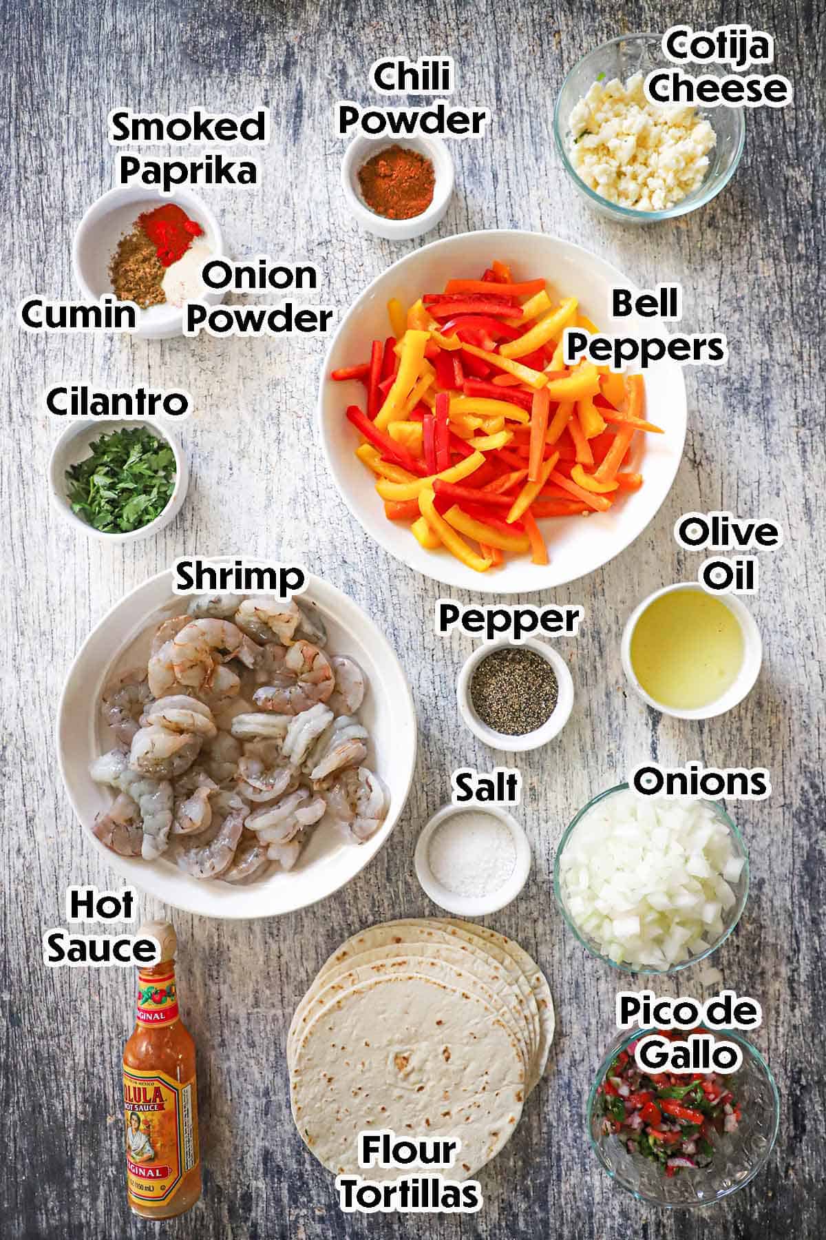An arrangement of ingredients for best shrimp fajitas on a grey wooden background including raw shrimp, red and yellow bell peppers, olive oil, flour flour tortillas, pico de gallo, cilantro, and seasonings.