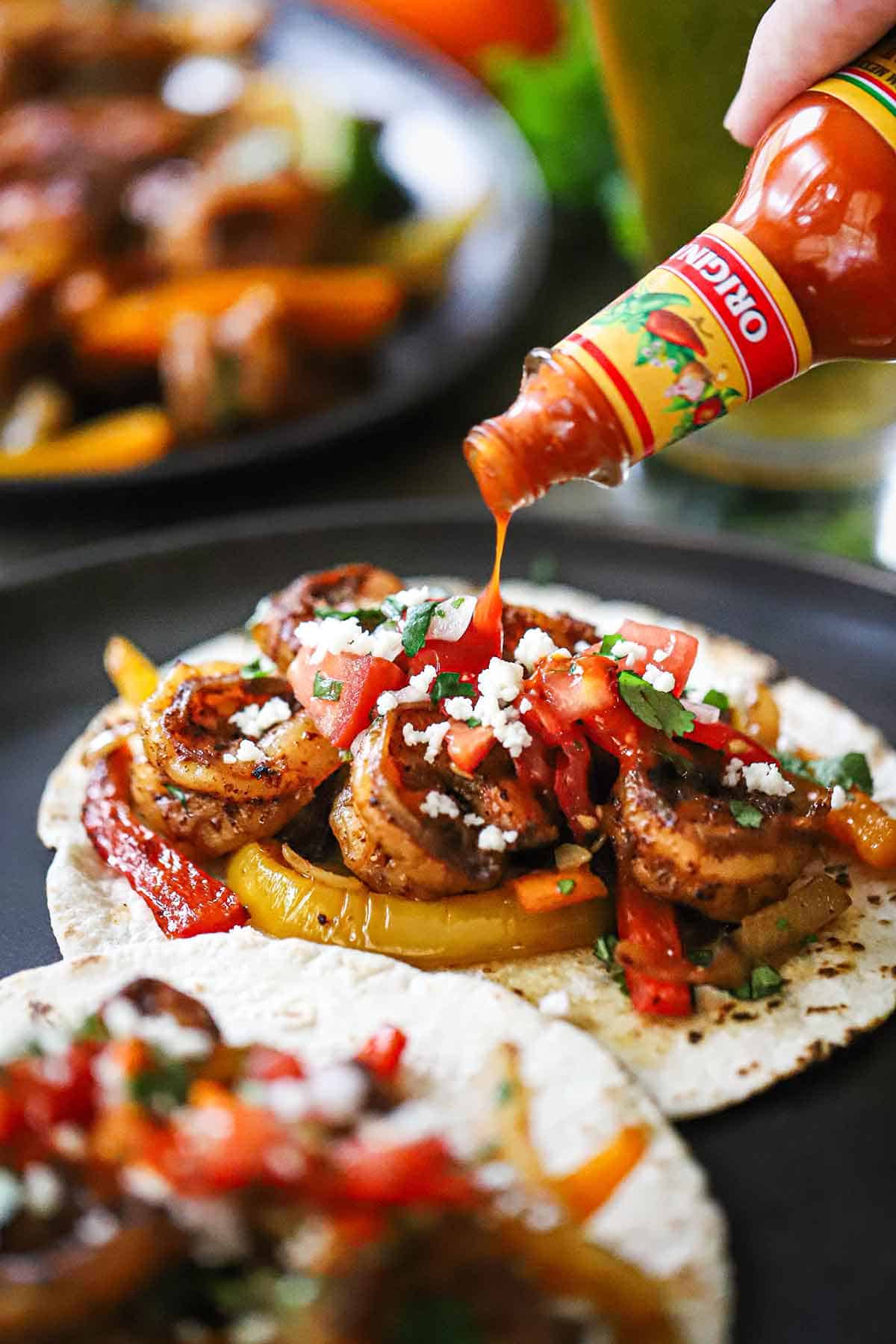 A person drizzling hot sour from a bottle over the top of an open shrimp fajita that is topped with chopped cilantro and crumbled cotija cheese.
