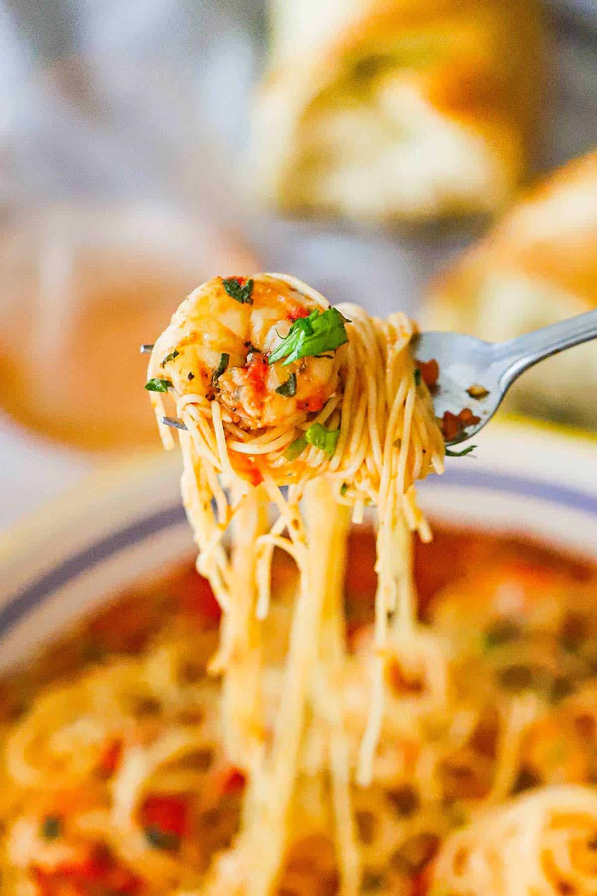 A person using a fork to raise a bite-full of angel hair pasta wrapped around the fork with a sautéed shrimp on the end of the fork.