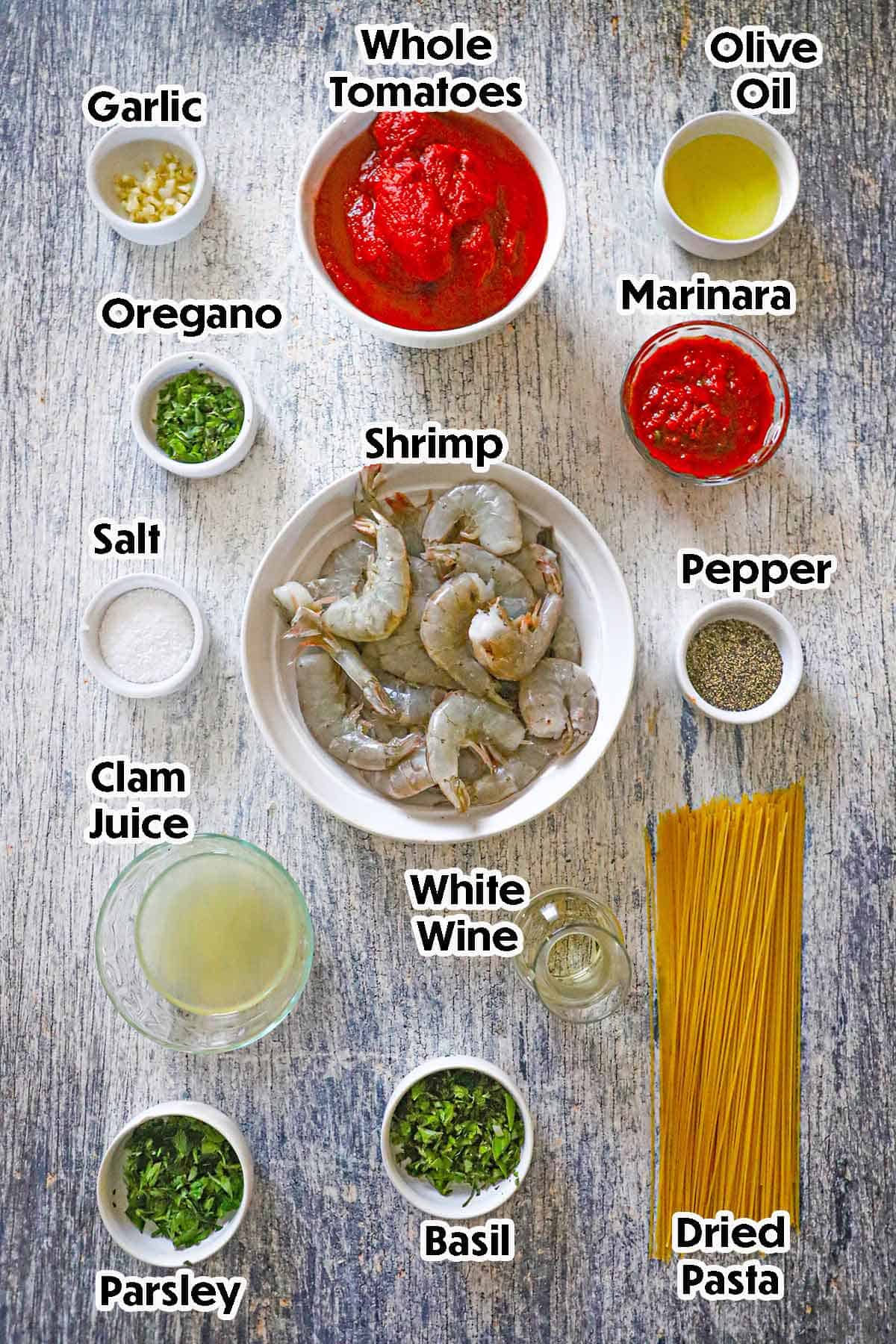 An arrangement of ingredients for shrimp marinara on a grey wooden background including raw shrimp, whole tomatoes, garlic, white wine, clam juice, olive oil, marinara sauce, dried pasta, seasonings, and fresh herbs.