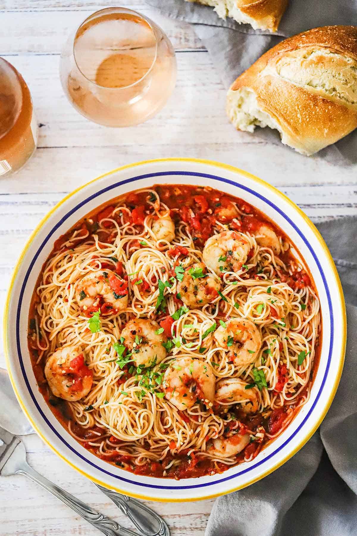 An overhead view of a large shallow pasta bowl filled with shrimp marinara and is garnished with chopped Italian parsley with a torn piece of Italian bread and two glasses of wine nearby.