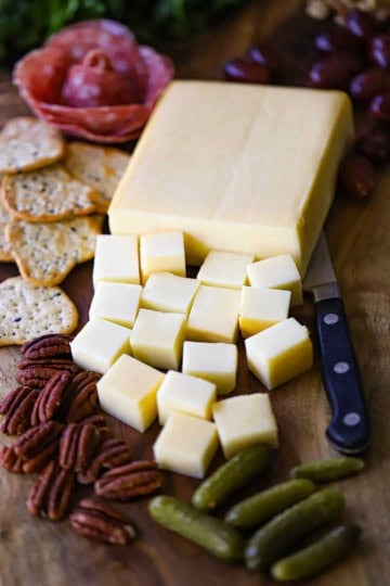 A straight-on view of smoked mozzarella that has partially been cut into cubes and is resting on a board next to crackers, pecans, salami, and baby dill pickles.