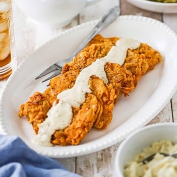 A close-up view of four Southern-Fried Pork Chops stacked against each other on an oval platter with a row of cream gravy across the top center of each chop.