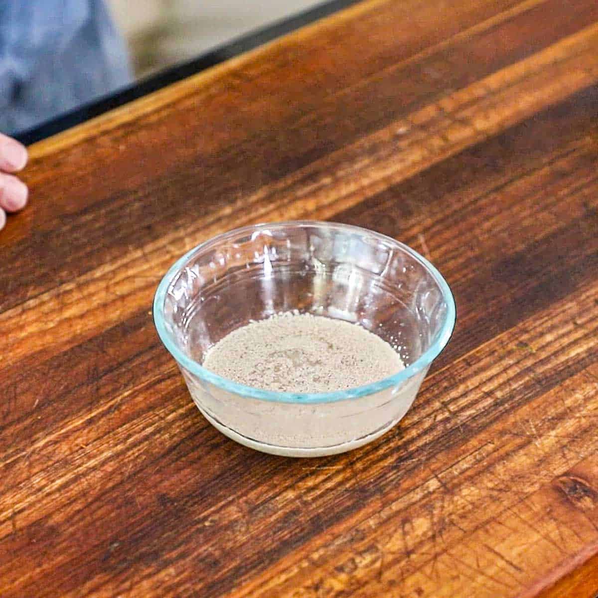 A small glass bowl partially filled with warm water that has active yeast blooming in it on a large wooden cutting board.