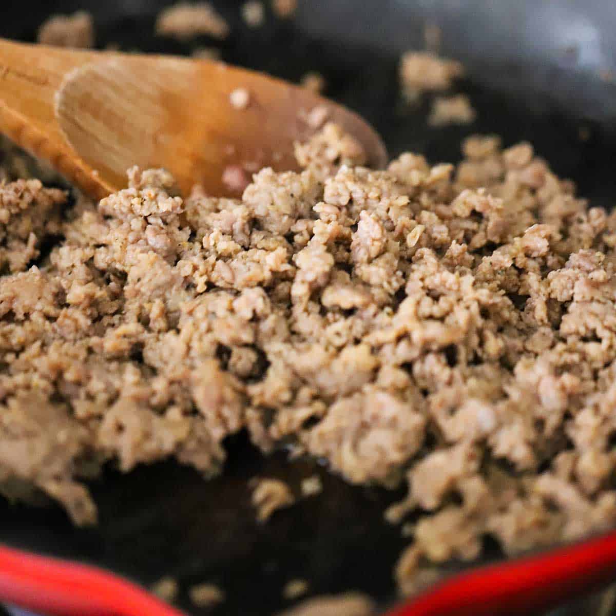 A close-up view of crumbled Italian sausage being cooked in a large ceramic skillet with a wooden spoon in the skillet.
