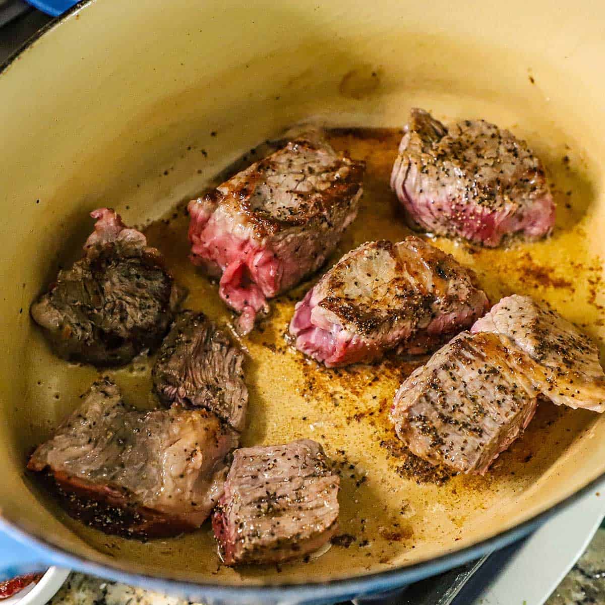 Eight chunks of seasoned beef chuck roast being seared in oil in a large Dutch oven.
