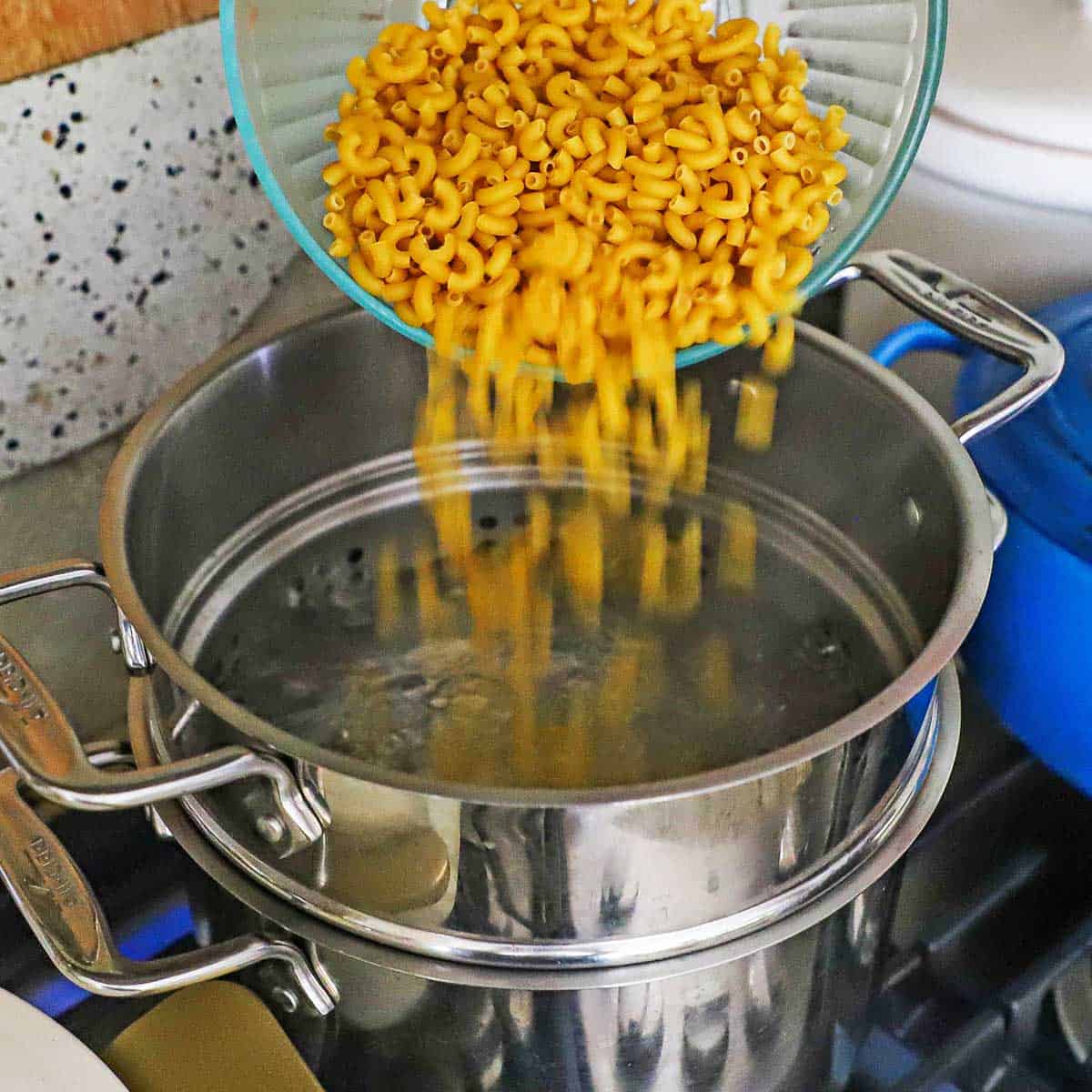 A person dumping uncooked macaroni into a pasta into pasta pot on a gas stove filled with boiling water.