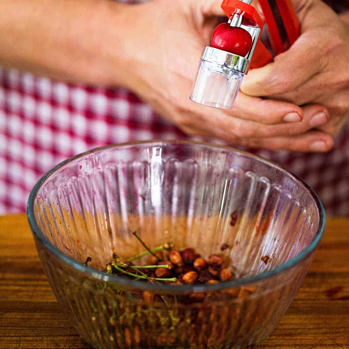 A person using a cherry pitter to remove the pit from a fresh bing cherry over a small glass bowl filled with more cherry pits and stems.