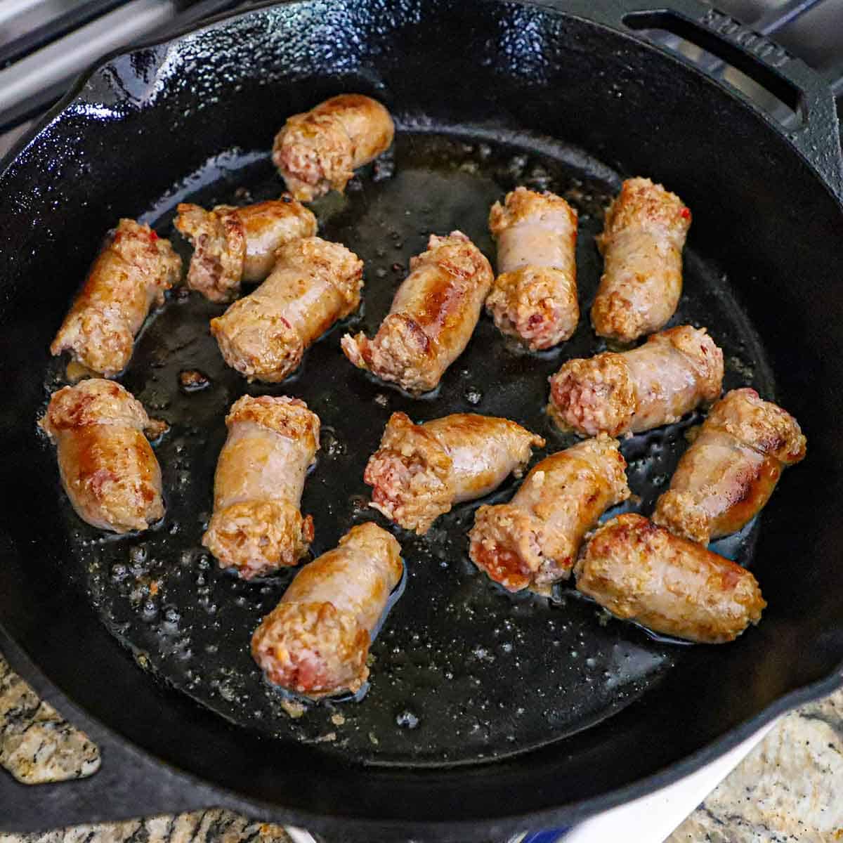 Two-inch strips of Italian sausage being seared in oil in a large cast-iron skillet on a gas stove.