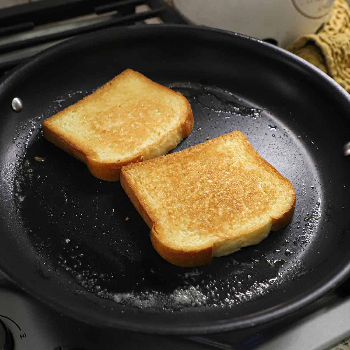 Two slices of brioche bread toasting in a large black non-stick skillet with melted butter in the pan.