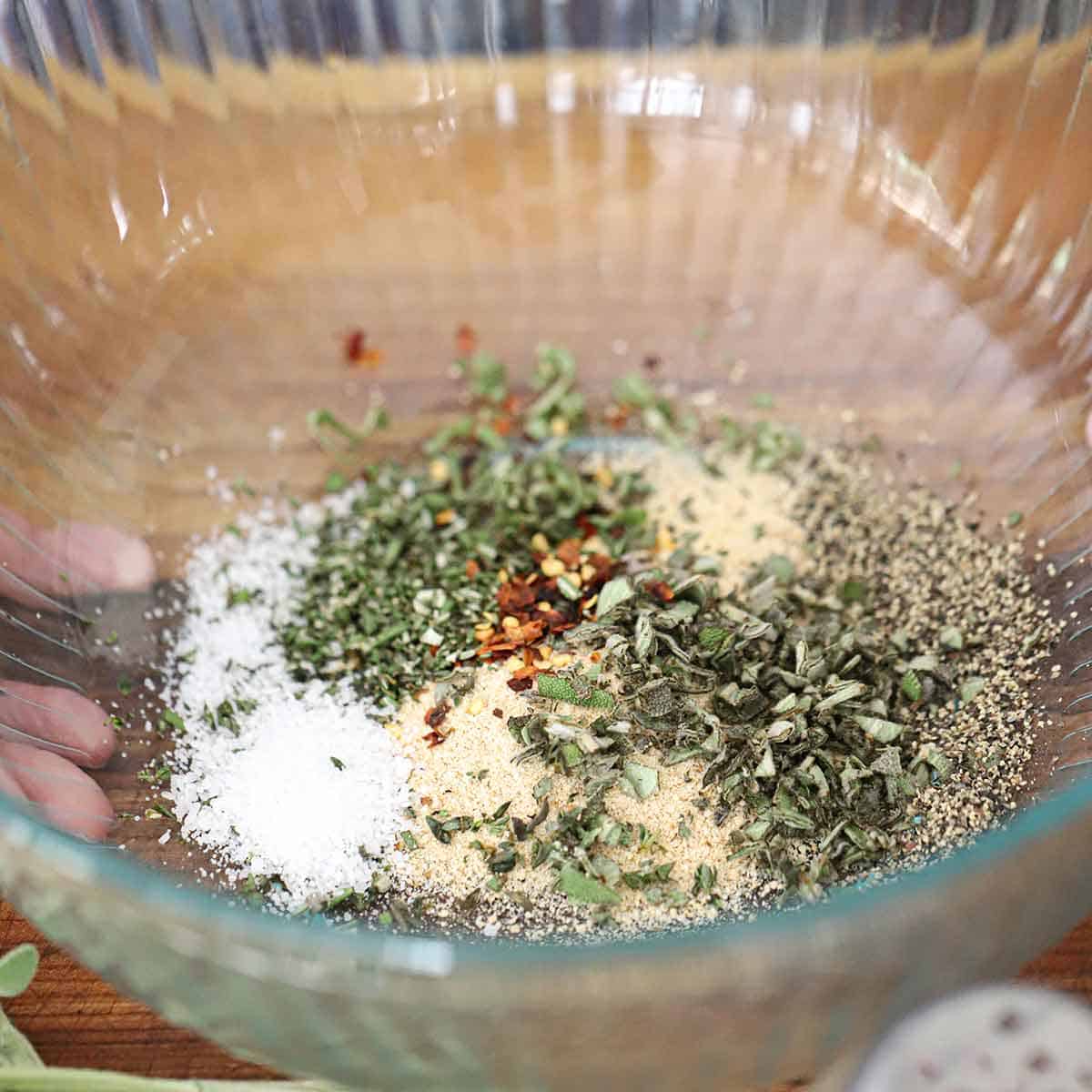 A large glass bowl filled with salt, breadcrumbs, red pepper flakes, sage, rosemary, and thyme.