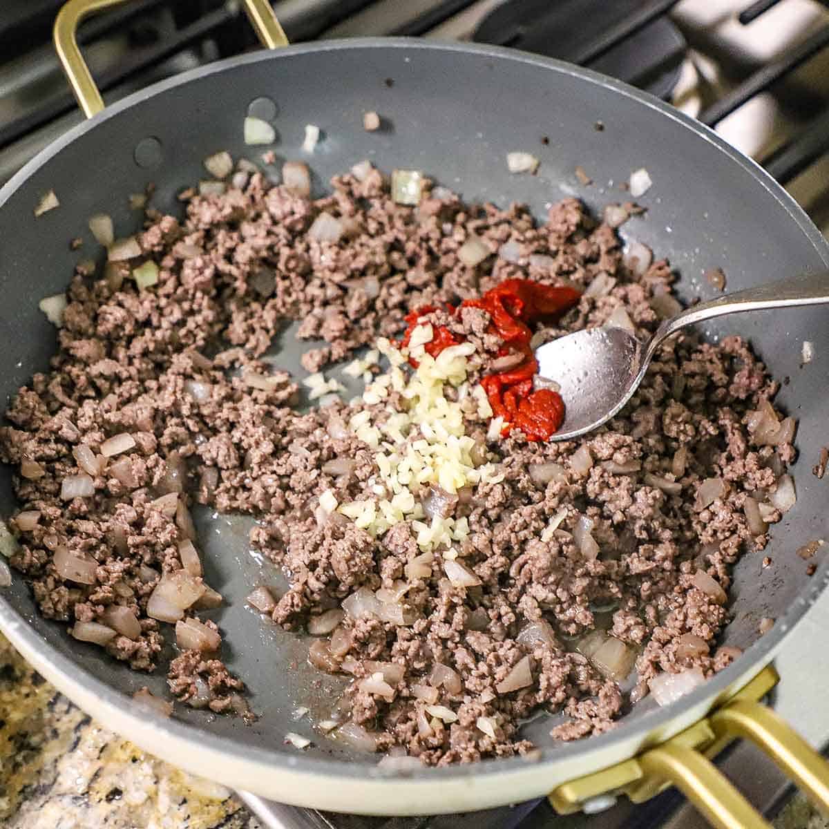 A person using a spoon to add tomato paste into a skillet that is filled with sautéed onions, minced garlic, and cooked ground beef on a gas stove.