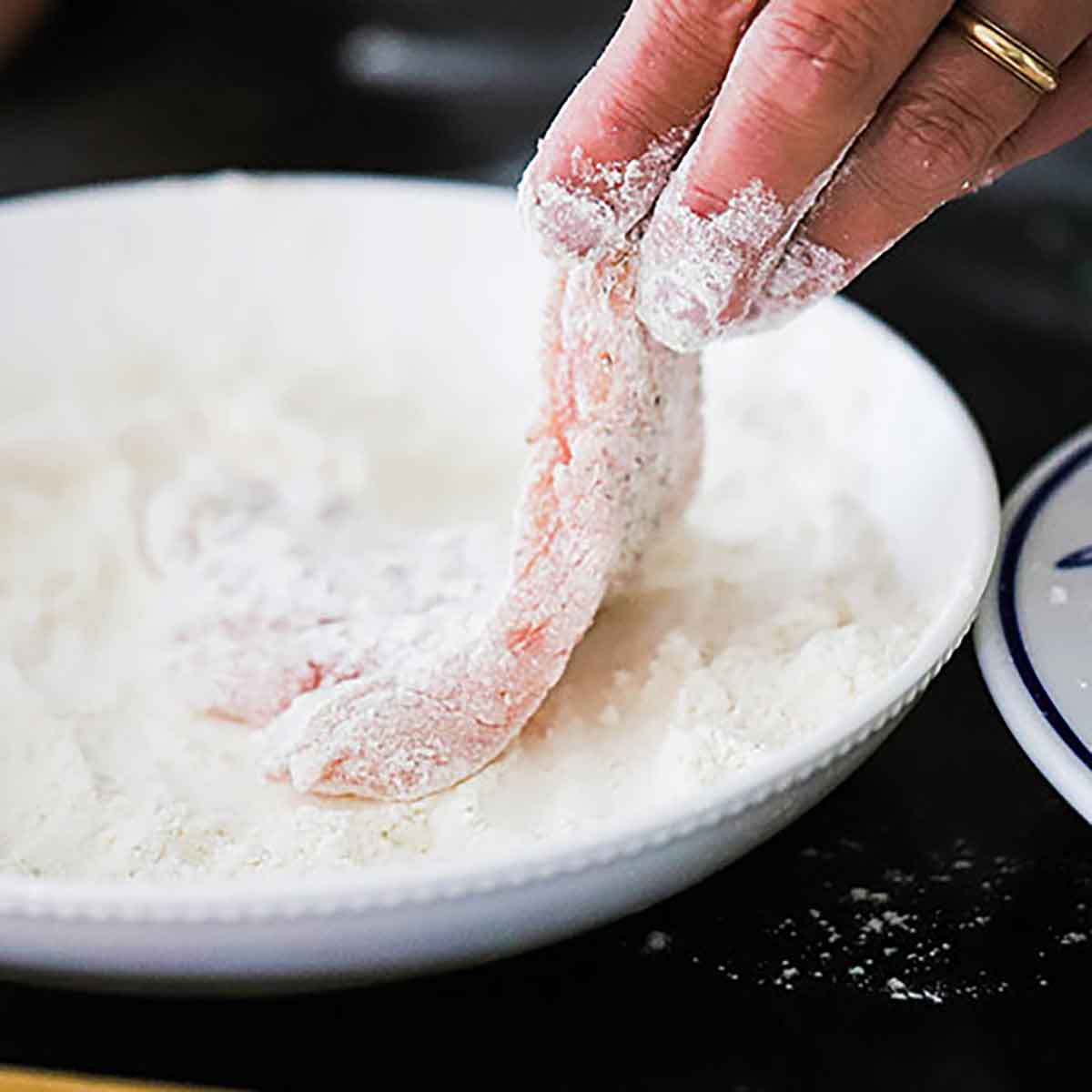 A person dredging a slice of pork tenderloin through a all-purpose flour in a shallow white bowl.