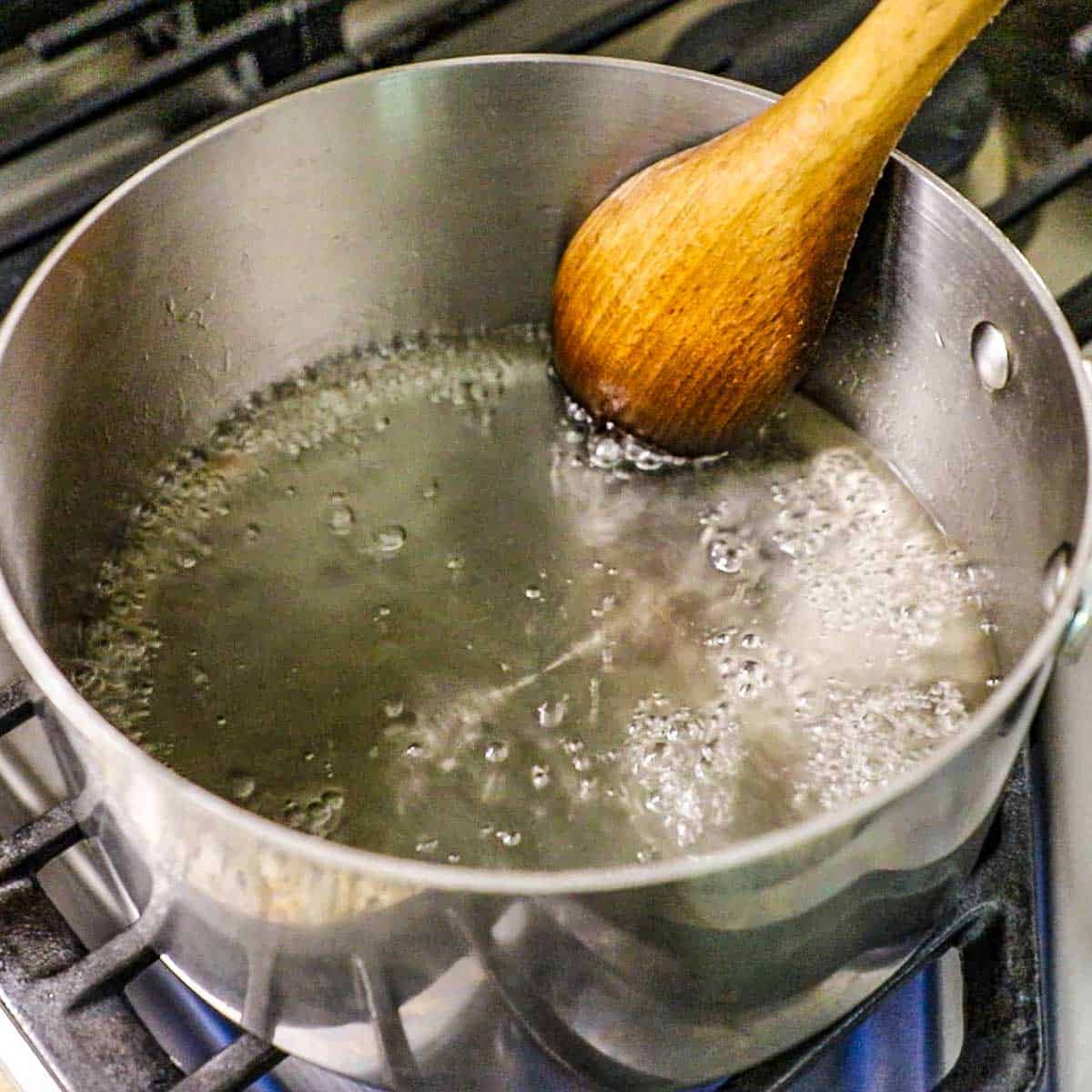 A person using a wooden spoon to stirring boiling water with dissolving sugar in it on a gas stove.