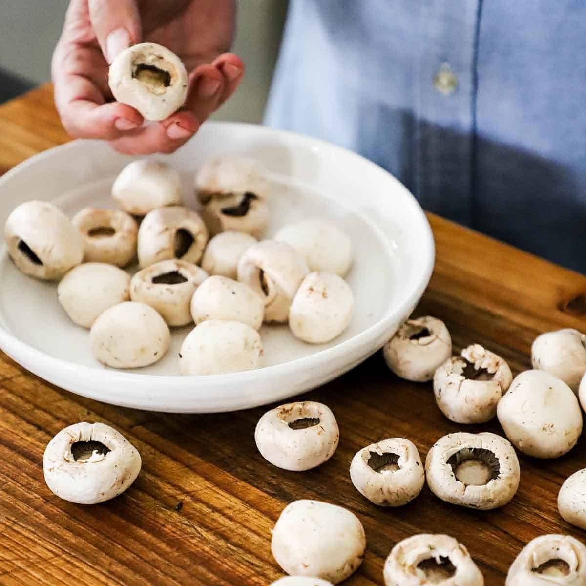 A person using his hand to remove the stems from white button mushrooms that are in a white bowl and also on a wooden cutting board.