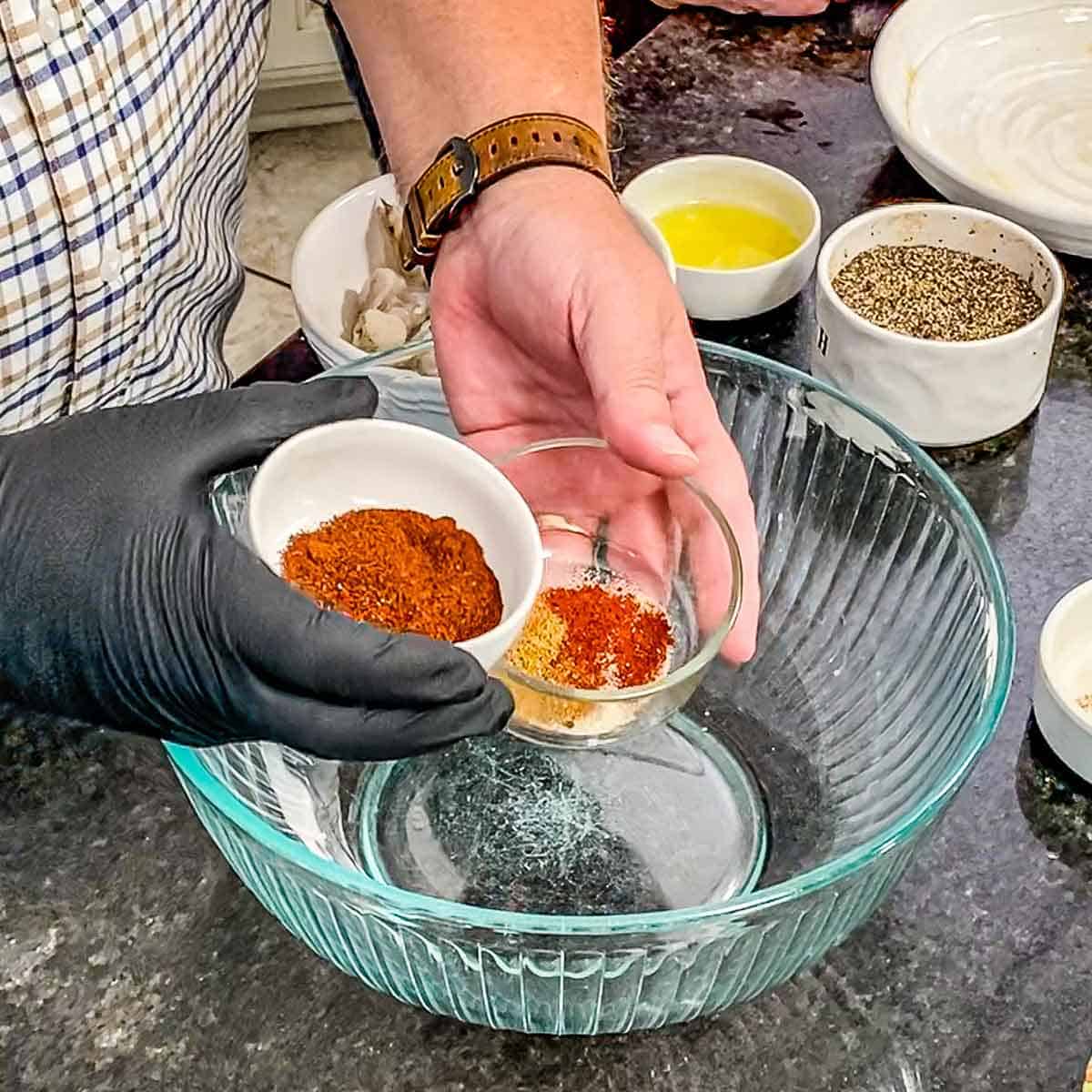 A person holding one small bowl of chili powder and ground cumin in one hand and another small bowl of paprika, onion powder, salt, and pepper in the other hand both over a large empty glass bowl.