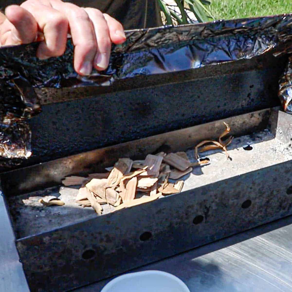A person lifting the cover of a electric smoker wood box revealing a few wood chips inside.