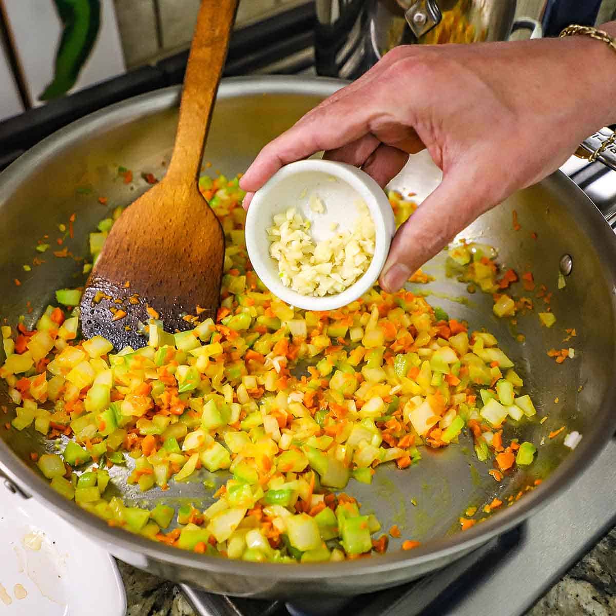 A person holding a small bowl containing minced garlic over a large silver skillet filled with sautéed onion, carrots, and celery.