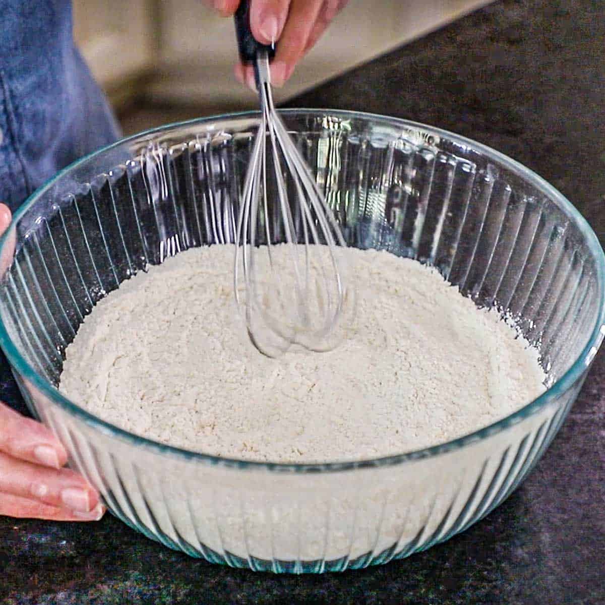 A person using a whisk to stir together a flour, salt, and baking powder in a large glass bowl.