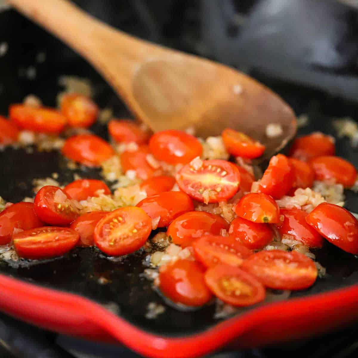 A close-up view of halved cherry tomatoes being sautéed with chopped shallots in butter in a large ceramic skillet.