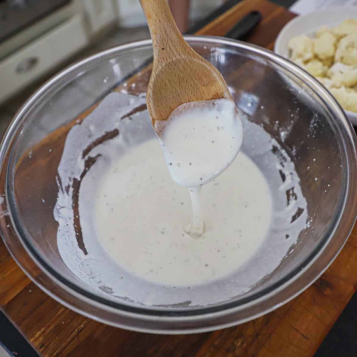 A person raising a large wooden spoon from a bowl filled with a white batter for deep-frying cauliflower florets.