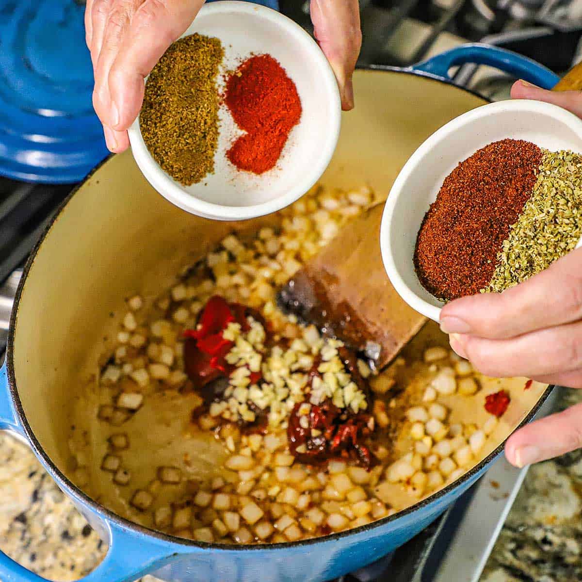 A person holding a small bowl filled with dried cumin and smoked paprika in one hand with the other hand holding a small bowl filled with chili powder and dried oregano both over a Dutch filled with with sautéed onions, garlic, and chipotle peppers.