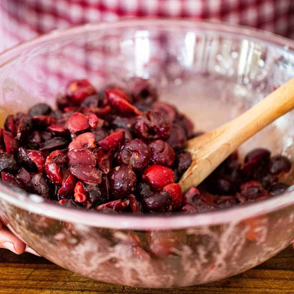 A person using a wooden spoon to stir pitted fresh cherries in a large glass bowl along with lemon juice, sugar, cornstarch, and vanilla extract.