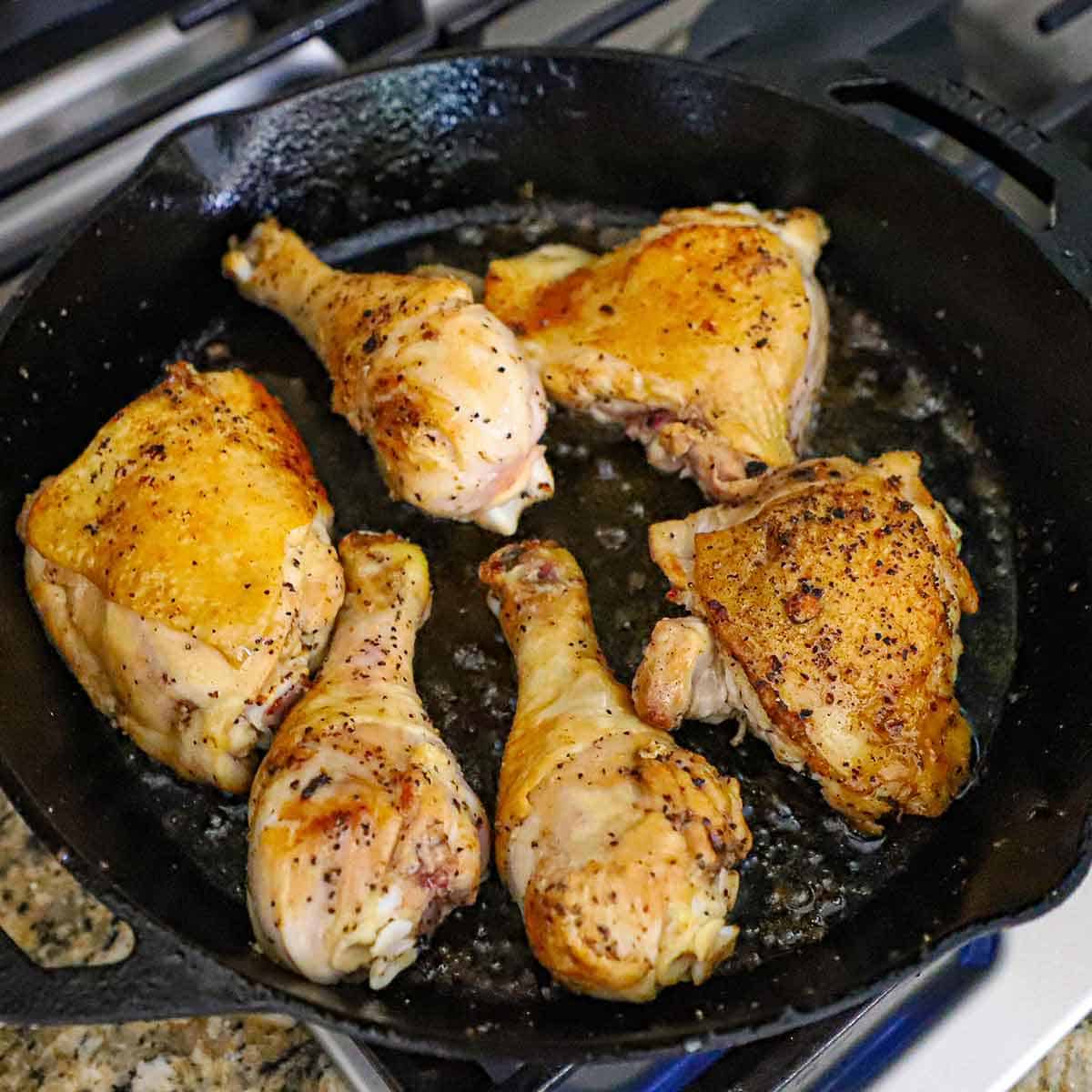 Three chicken legs and three chicken thighs being seared in oil in a large cast-iron skillet on a gas stove.