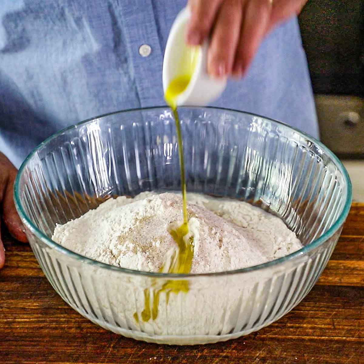 A person pouring olive oil from a small bowl into a large glass bowl filled with flour, fine sea salt, sugar, and instant dry yeast.