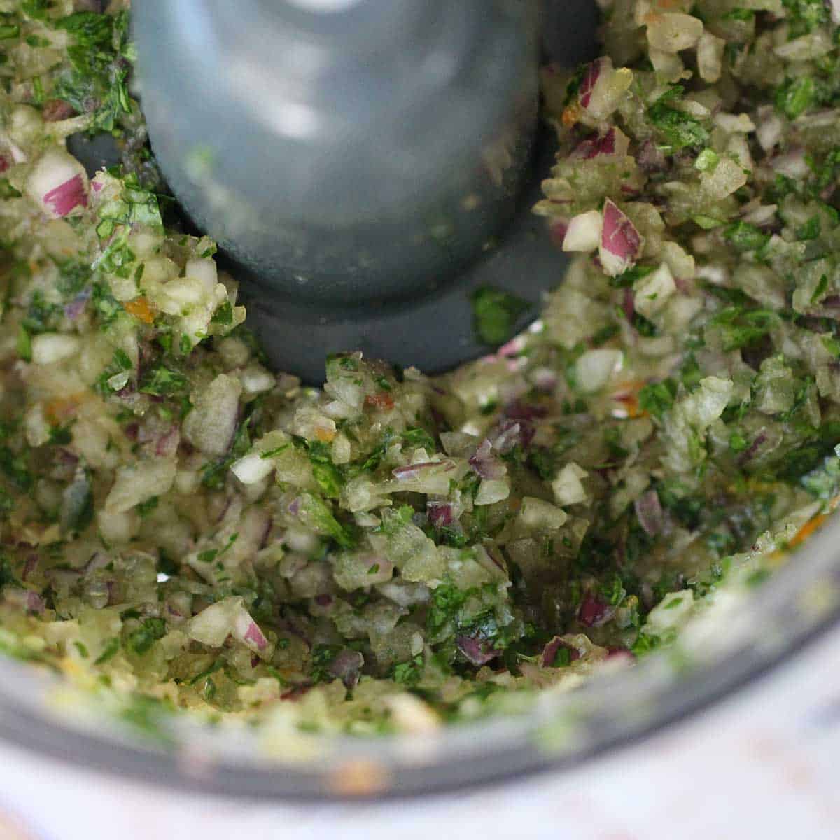 A close-up view of finely chopped Italian parsley and onion in the bowl of a food processor.