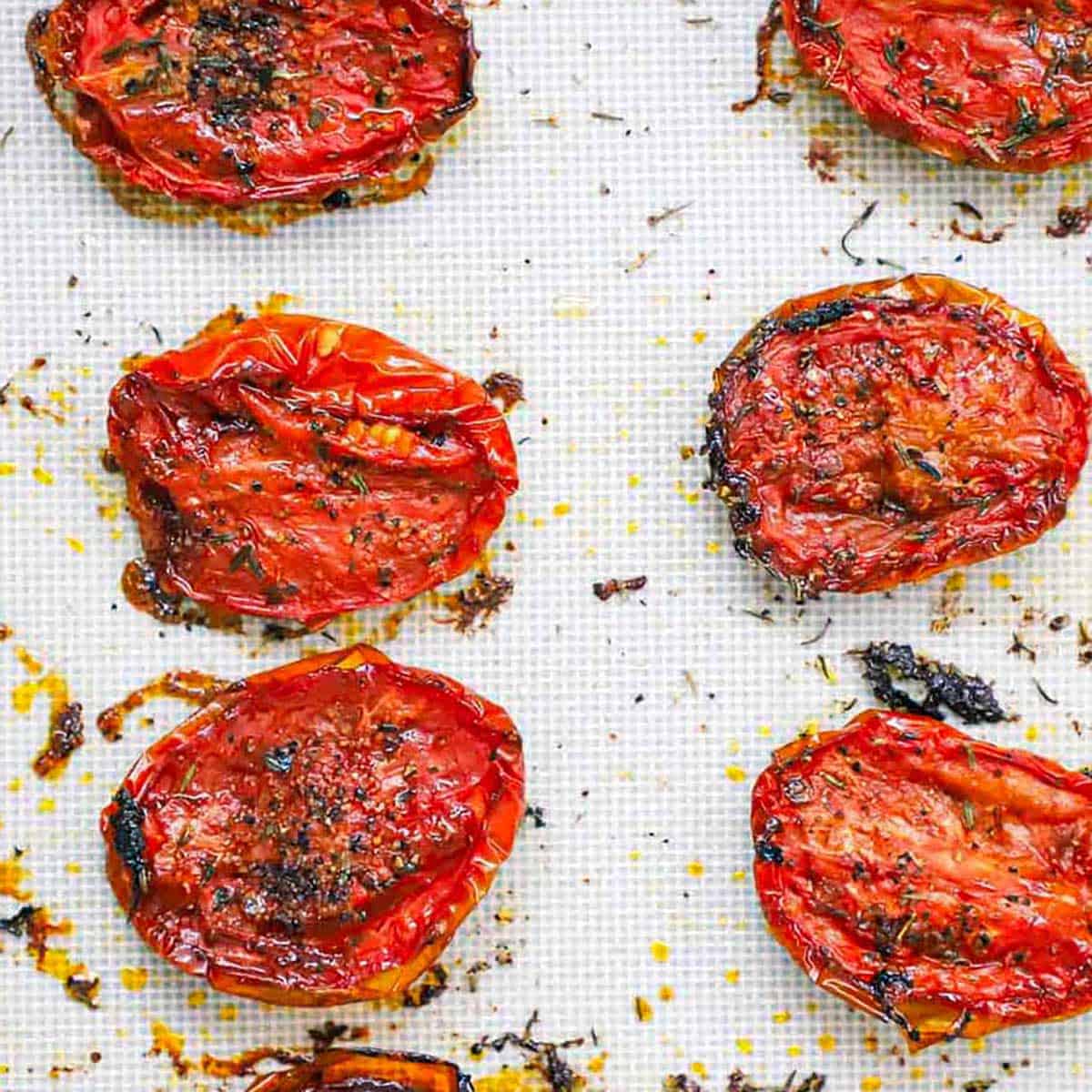An overhead view of two roasted of roasted sliced tomatoes that have been seasoned with salt, pepper, and thyme resting on a silicone mat.