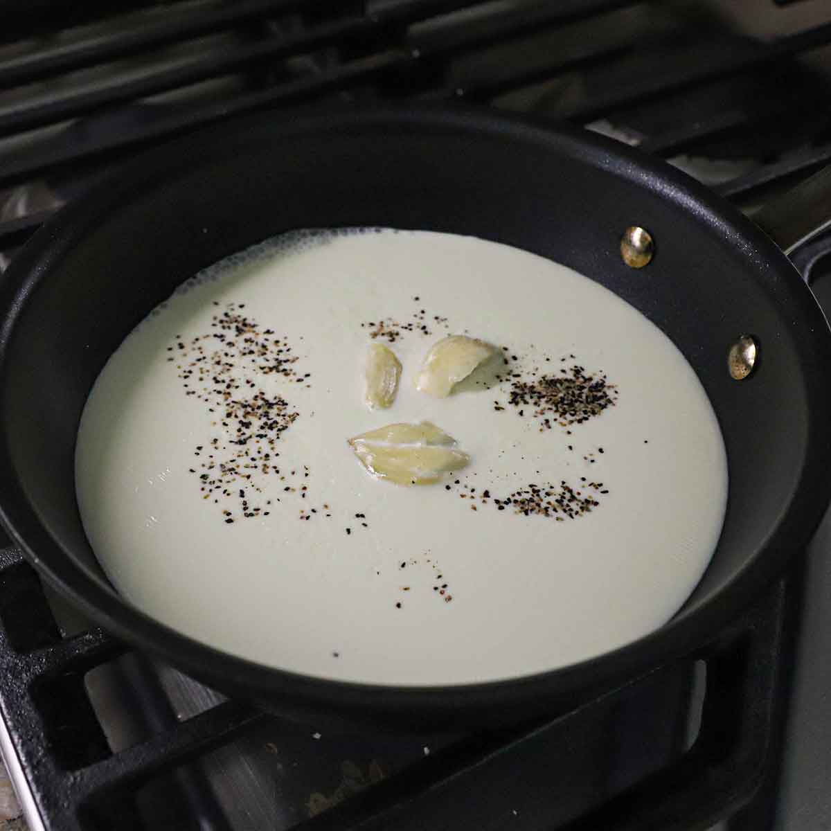 A small black non-stick skillet partially filled with heavy cream, three smashed cloves of garlic, salt, and cracked black pepper.